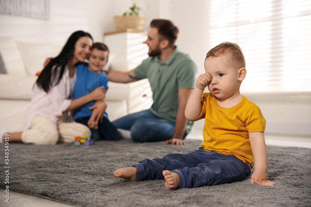 Unhappy baby sitting alone on floor while parents spending time with his elder brother at home. Jealousy in family Unhappy baby sitting alone on floor while parents spending time with his elder brother at home. Jealousy in family