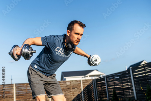 Get in shape without gym. A young guy in sportswear is training in the backyard, he is doing arm and shoulder exercise with iron dumbbells. Concept of healthlife, sport, selfdissipline