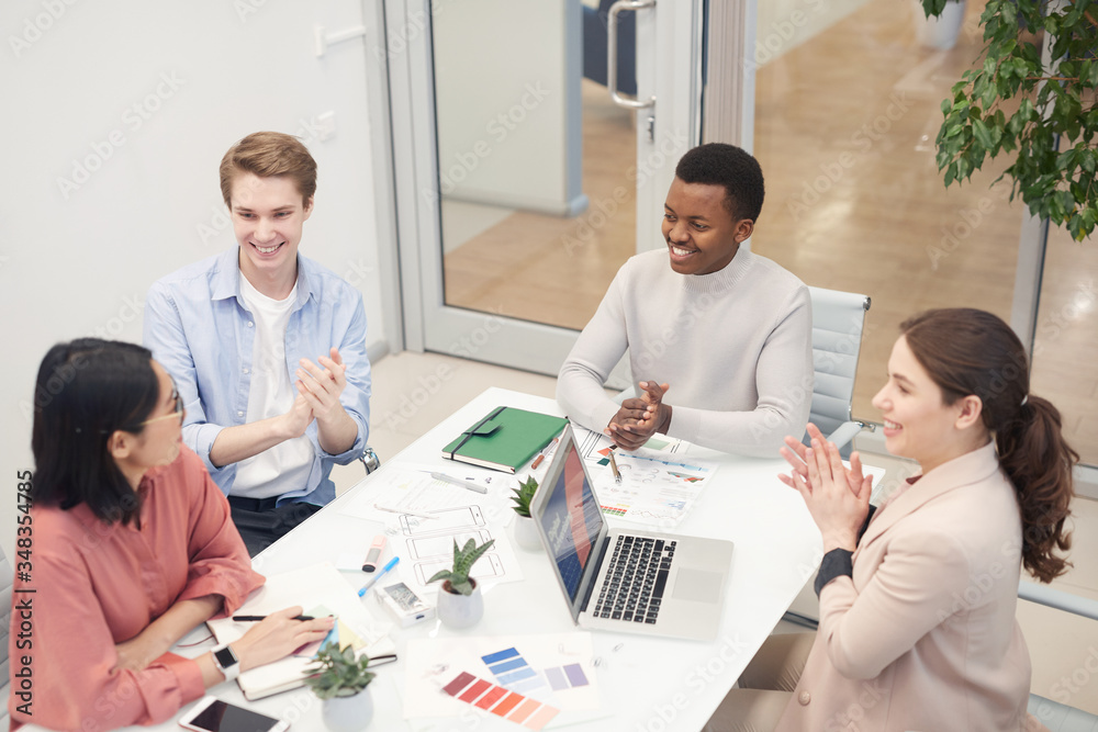 High angle view at multi-ethnic group of business people clapping to Asian colleague while collaborating on design project during meeting in office, copy space High angle view at multi-ethnic group of business people clapping to Asian colleague while collaborating on design project during meeting in office, copy space