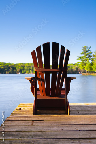 Muskoka chair sitting on a wood dock facing a lake. Across the calm water is a cottage nestled among green trees.