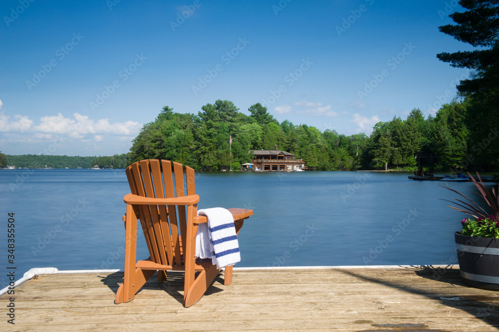 Muskoka chair sitting on a wood dock facing a calm lake. Across the water is a cottage nestled among green trees. A towel is folded on the chair arm.