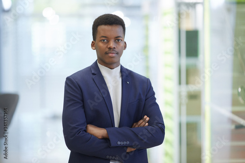 Waist up portrait of successful African businessman posing confidently while standing with arms crossed in modern office interior, copy space