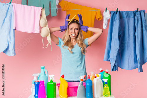 Sad housewife holds head with hands, in front of her is basin and cleaning supplies, behind is clean clothes on rope