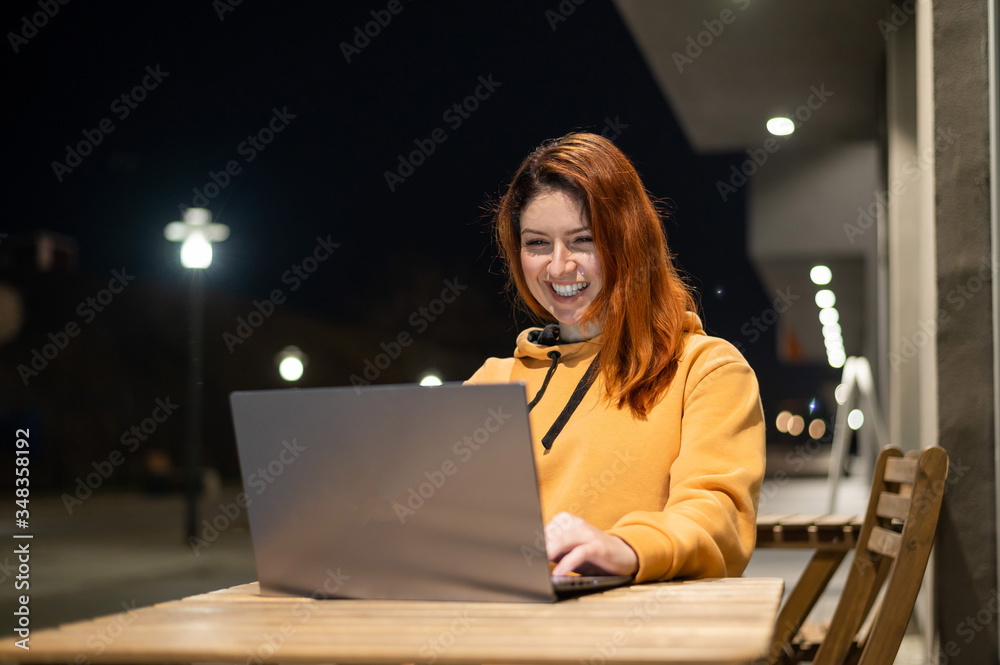 A woman works remotely at a laptop in a summer cafe late in the evening. Happy Girl studying while sitting on an empty street at a wooden table. Female freelancer in a sweatshirt at the evening A woman works remotely at a laptop in a summer cafe late in the evening. Happy Girl studying while sitting on an empty street at a wooden table. Female freelancer in a sweatshirt at the evening