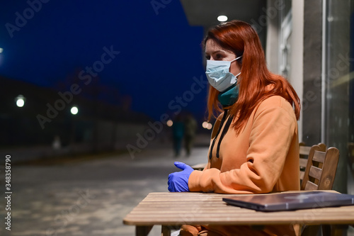 A woman in a medical mask and gloves walks late at night alone. The girl is sitting at a wooden table with a laptop in quarantine. Coronavirus.