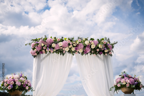 against the blue sky stands a wedding arch decorated with flowers, two vases with flowers stand on white pedestals, festive decor on a vadba, wedding of the newlyweds