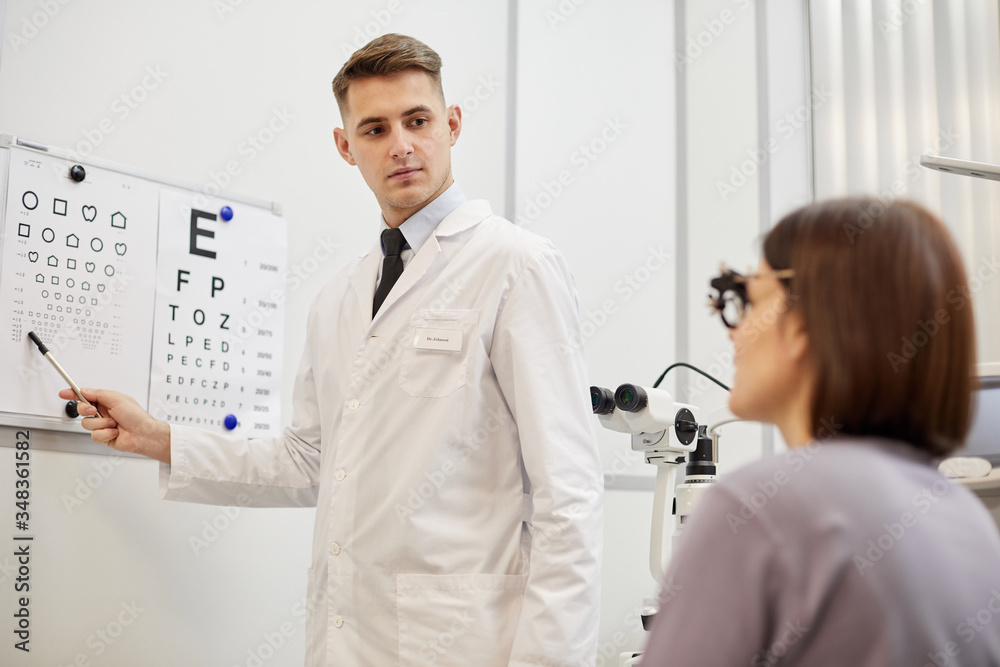 Obraz premium Portrait of young optometrist pointing at vision chart while checking eyesight of female patient in modern ophthalmology clinic