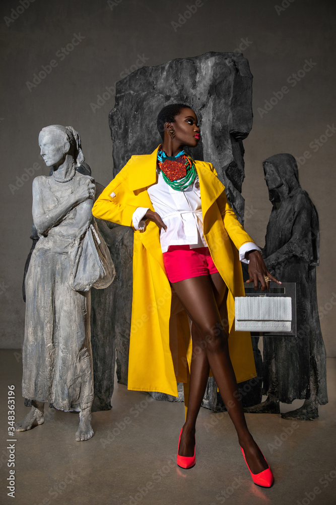 An elegant young black woman with short black hair & beautiful makeup and gorgeous jewelry posing by herself indoors in front of statues is wearing a yellow coat, white blouse & red shorts.