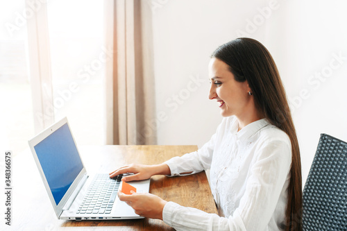 A woman using laptop for online shopping, online banking. She sits at the table with a credit card in hand and looks at the screen with a smile