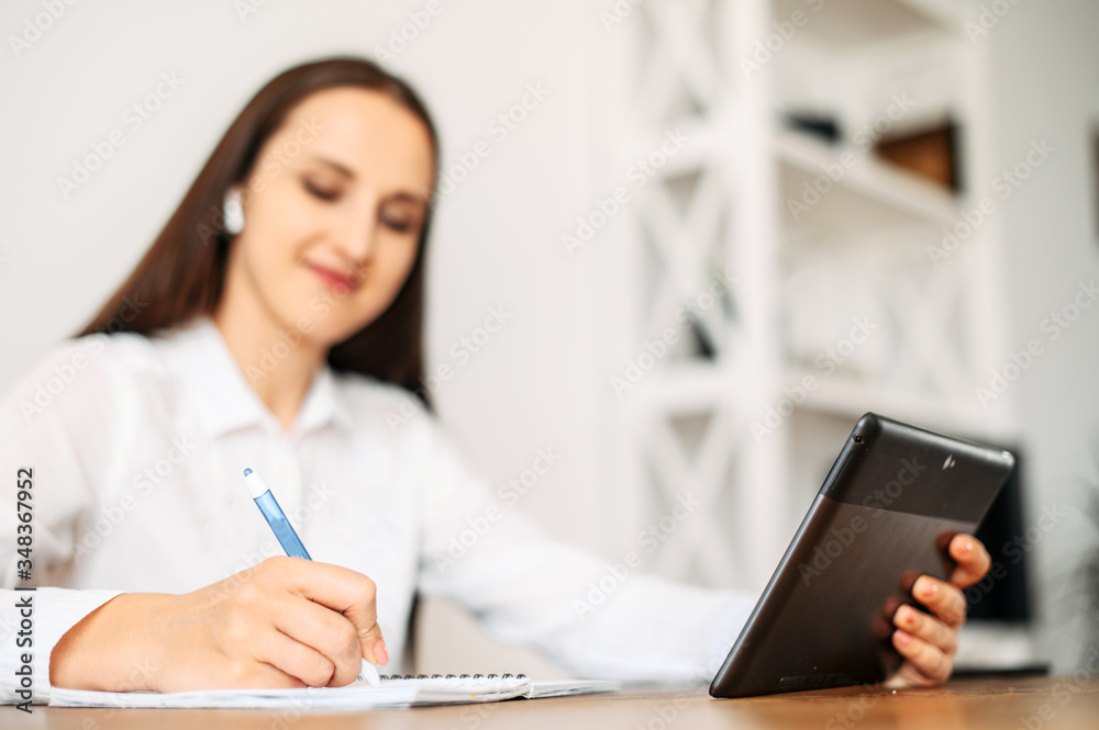 Online learning online, online classes. A female student is using tablet for watching online lectures, she is doing notes in notebook while sitting at the table