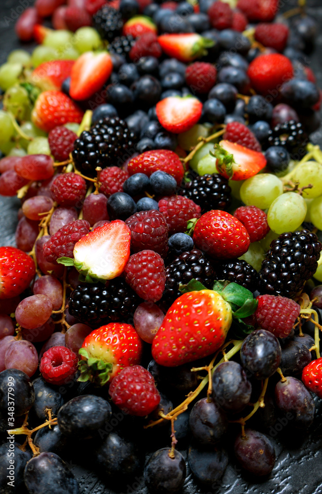 Obraz premium Summer berries food background full frame close up with strawberries, blackberries, blueberries, red, dark and green grapes on black marble.