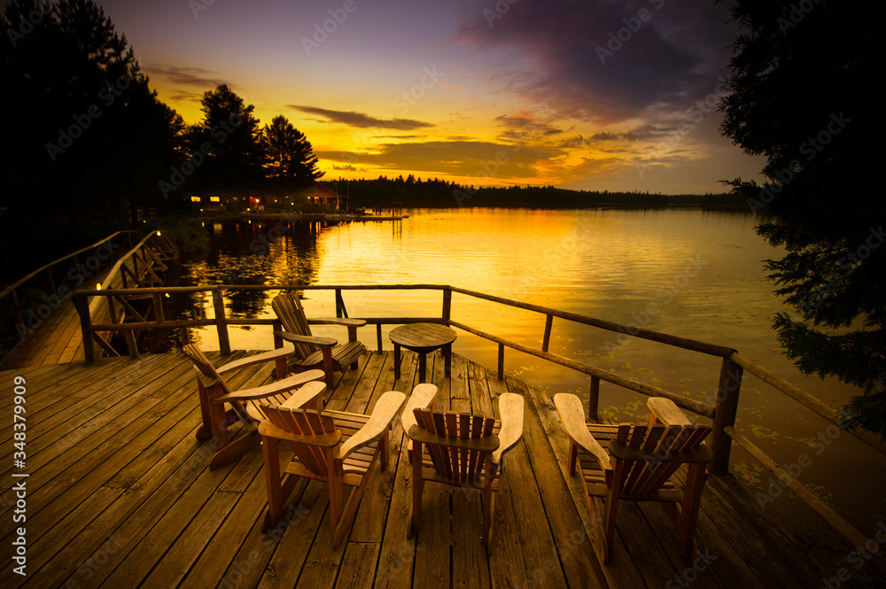 Adirondack chairs sitting on a wooden dock facing a lake at sunset. Adirondack chairs sitting on a wooden dock facing a lake at sunset.