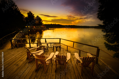 Adirondack chairs sitting on a wooden dock facing a lake at sunset.