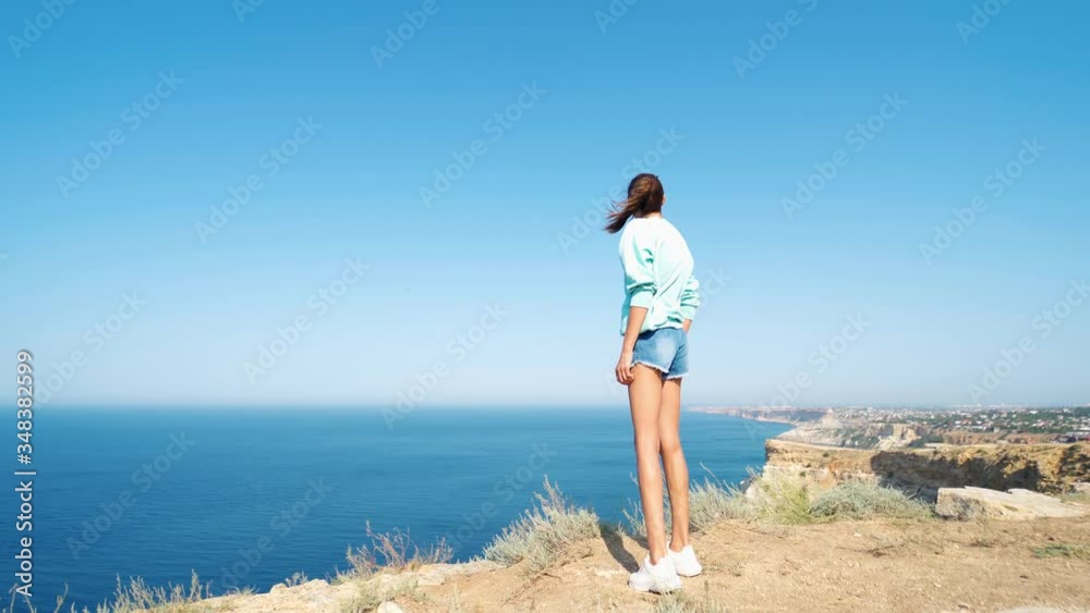 back view of carefree young woman standing with raised hands on cliff edge with beautiful sea view and enjoying live nature. female tourist having great time on vacation. Full length 4k footage back view of carefree young woman standing with raised hands on cliff edge with beautiful sea view and enjoying live nature. female tourist having great time on vacation. Full length 4k footage