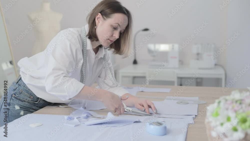 Slow motion video of the fashion designer works at a table with curves and a pattern, she are cutting fabric, around lies scissors, sew machine and dummy on the background, camera is moving back Slow motion video of the fashion designer works at a table with curves and a pattern, she are cutting fabric, around lies scissors, sew machine and dummy on the background, camera is moving back