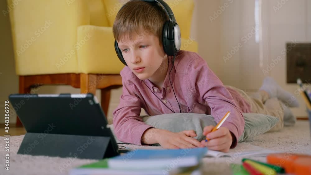 smart school boy pupil looks on tablet screen and writes homework in a notebook. Spbas boy lying on floor stay home and study at distant learning, quarantine. concentrated child listen to teacher smart school boy pupil looks on tablet screen and writes homework in a notebook. Spbas boy lying on floor stay home and study at distant learning, quarantine. concentrated child listen to teacher