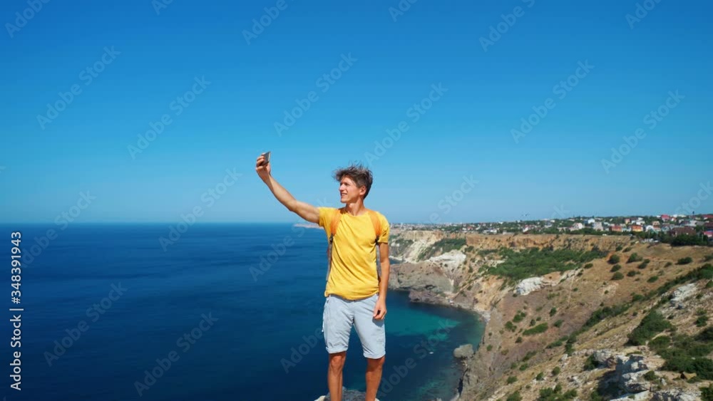 young male tourist making selfie and smile to the camera on cliff edge with beautiful sea view and enjoying wonderful nature. Caucasian man having a great time on his vacation.