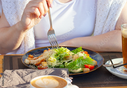 Fototapeta Naklejka Na Ścianę i Meble -  Caesar salad on a dark plate and the hands of a girl in a white T-shirt, which is going to eat a salad, with a fork. A plate on the table and two coffees.