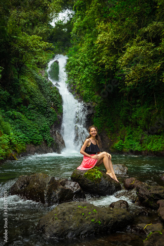 Happy smiling Caucasian woman sitting on the rock and enjoiyng waterfall landscape. Travel concept. Dedari waterfall in Sambangan  Bali.