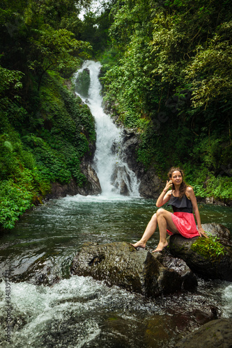 Attractive Caucasian woman sitting on the rock and enjoiyng waterfall landscape. Travel lifestyle. Dedari waterfall in Sambangan  Bali.