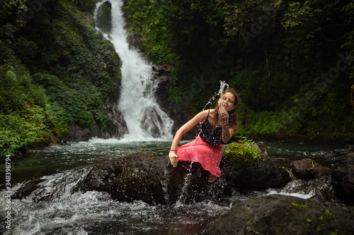 Young Caucasian woman sitting on the rock and playing with water in the river near the waterfall. Travel lifestyle. Dedari waterfall in Sambangan, Bali.