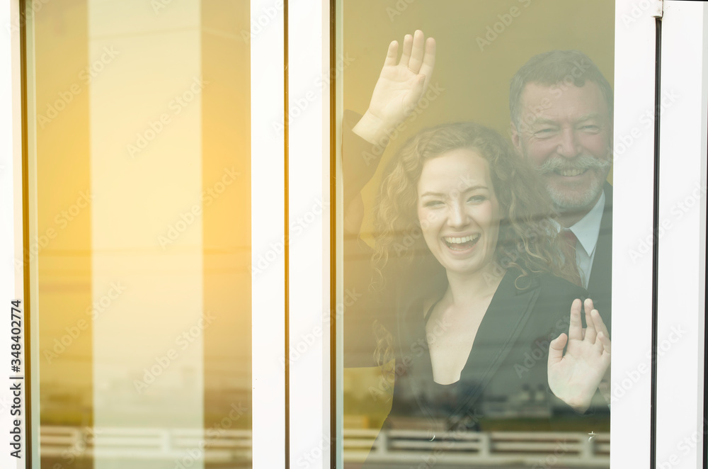 Business middle aged man and beautiful woman behind their glass window of home say hello to others people,Happy old man and young woman in suit standing and gesturing raised up hands.Work form home. Business middle aged man and beautiful woman behind their glass window of home say hello to others people,Happy old man and young woman in suit standing and gesturing raised up hands.Work form home.