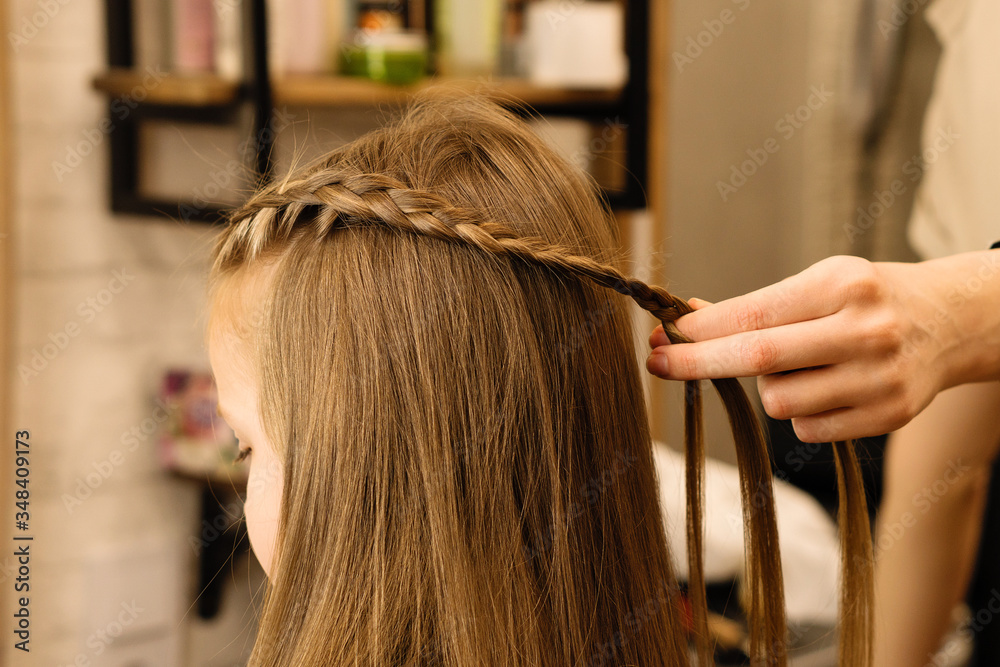 Naklejka premium Process of braiding the master weaves braids on her head blond little girl in beauty salon close up. Professional hair care and creating hairstyles.