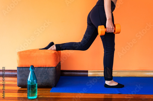 Legs of a slim young woman doing a bulgarian split squat, with orange dumbbells, leaning on a piece of furniture