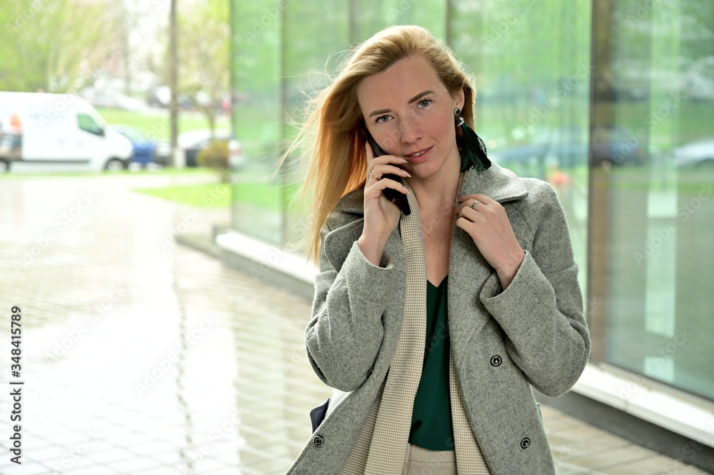 Photo portrait of a young business woman blonde smiling posing talking on the phone on a background of a city outdoors Photo portrait of a young business woman blonde smiling posing talking on the phone on a background of a city outdoors