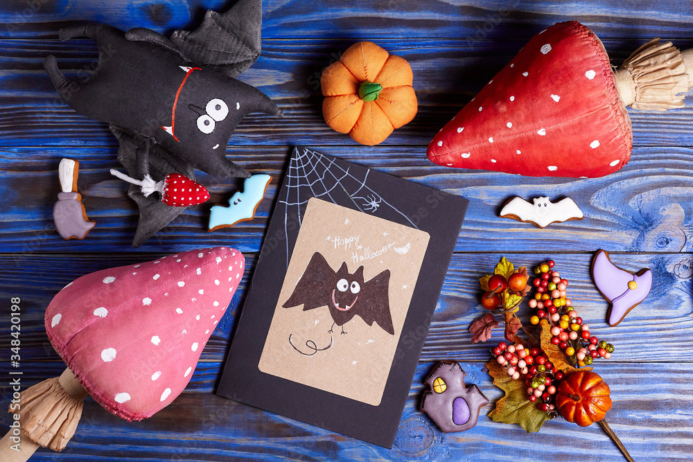 Halloween card with bat, bat textile toy, pumpkins, fly agaric and gingerbread cookies on blue wooden background