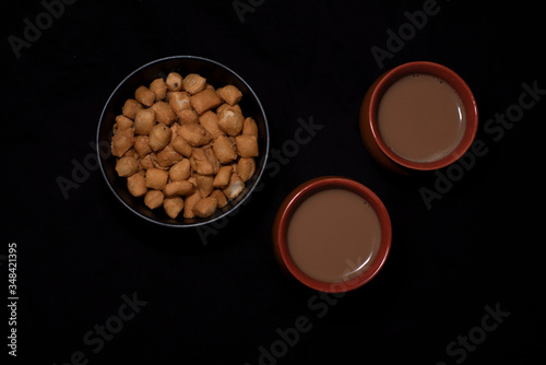 Top down image of infused tea in earthen cups along with a bowl of snacks kept in black copy space background. Indian beverages and food photography.