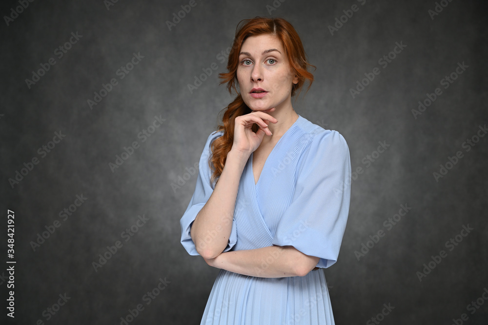 Pretty model posing with different emotions on a gray background in the studio. Portrait of a young caucasian woman with long red hair in a blue dress.