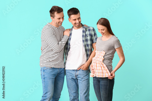 Gay couple and pregnant woman with baby clothes on color background