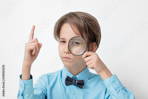a boy in a blue shirt and bow tie looks through a magnifying glass, serious smart boy, hand with the index finger pointing up , concept of knowledge, science study, research