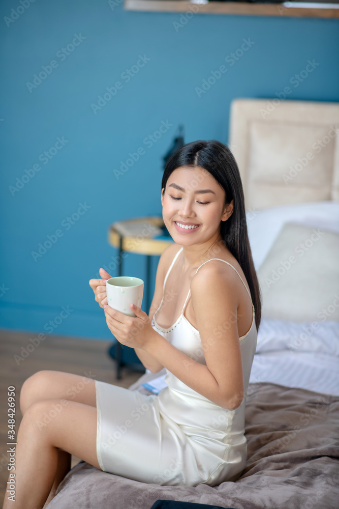 Young brunette in night dress sitting on bed enjoying her morning coffee Young brunette in night dress sitting on bed enjoying her morning coffee