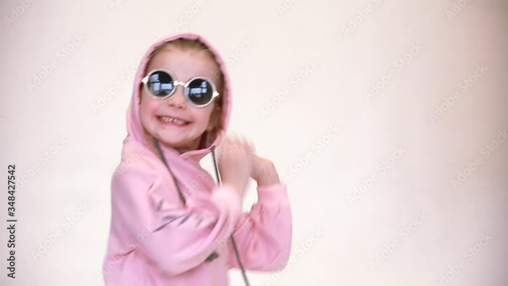 Fashionable girl in a pink bike with a hood and black sunglasses dancing on a gray-white background. 
