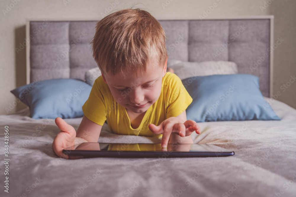 Distance education, learning. Boy is lying on the bed with a tablet in the room. Child with a gadget. Teenage child using tablet pc. Distance education, learning. Boy is lying on the bed with a tablet in the room. Child with a gadget. Teenage child using tablet pc.