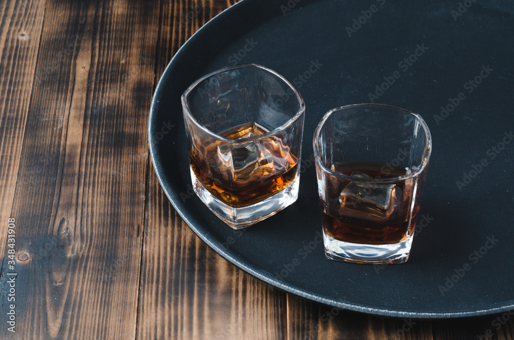Glasses of whiskey with ice cubes on a black tray and wooden table/Two Glasses of whiskey with ice cubes on a black tray and wooden table with copyspace. Glasses of whiskey with ice cubes on a black tray and wooden table/Two Glasses of whiskey with ice cubes on a black tray and wooden table with copyspace.