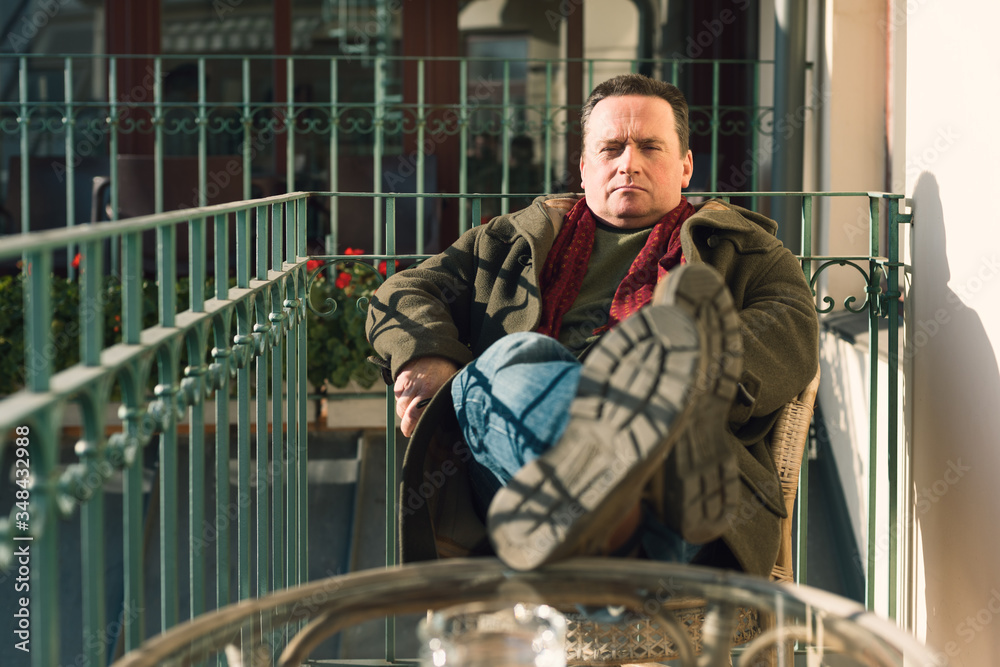 Middle-aged rich European man sitting on his balcony in a city, resting and basking in the sun, with his legs on the table. Concept of boredom at home Middle-aged rich European man sitting on his balcony in a city, resting and basking in the sun, with his legs on the table. Concept of boredom at home