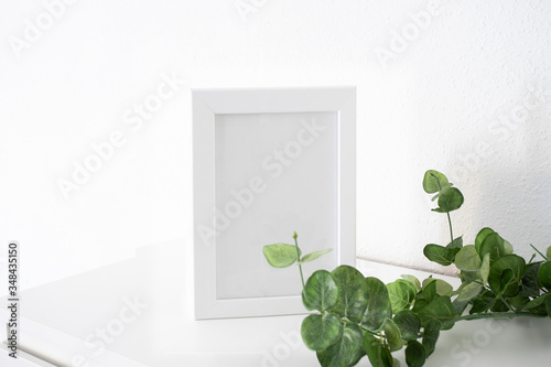 White frame mockup with green plant on a white table against white wall. Empty poster frame mockup for design.