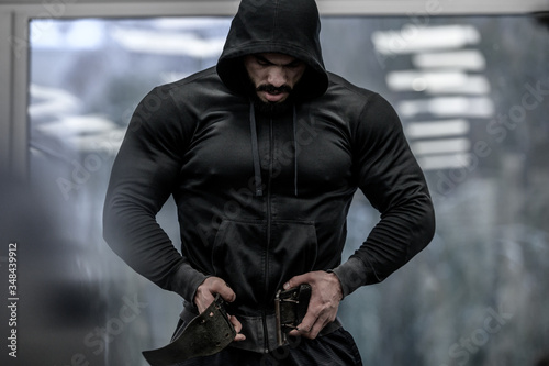 strong young bearded mighty man in hoodie wearing powerlifting belt for heavy hardcore weight training workout indoors sport fitness gym photo