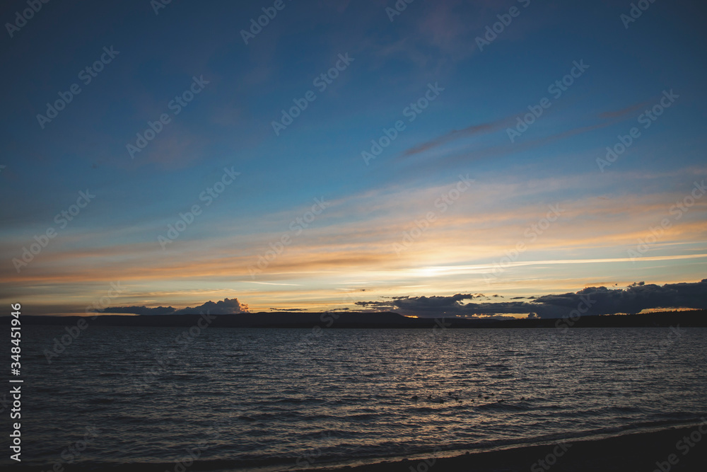 Sunset in orange yellow shades on blue sky and lake on the bottom with small clouds. Horizon travel wallpaper with nature concept and coastline there. Yellowstone lake in national park United States.