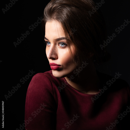 Charming young woman with blue eyes and long fair hair posing at studio over dark background. Amazing stylish girl with perfect makeup wearing elegant deep red dress. Beauty and fashion
