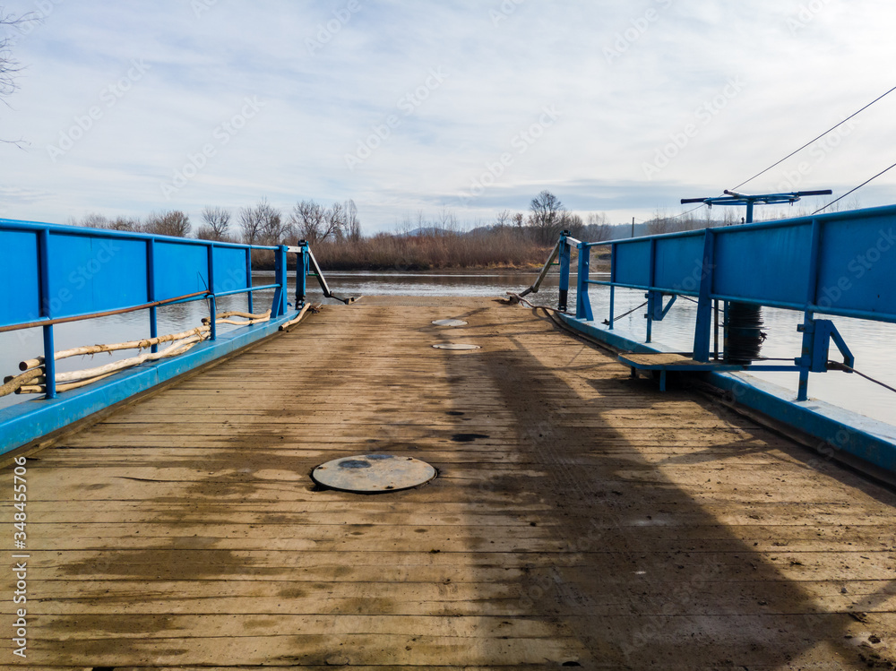 Obraz premium Ferry for transporting goods and people across the river moored along the river bank with a lowered ramp ready for loading. Raft made of metal with metal fences and wooden floor.