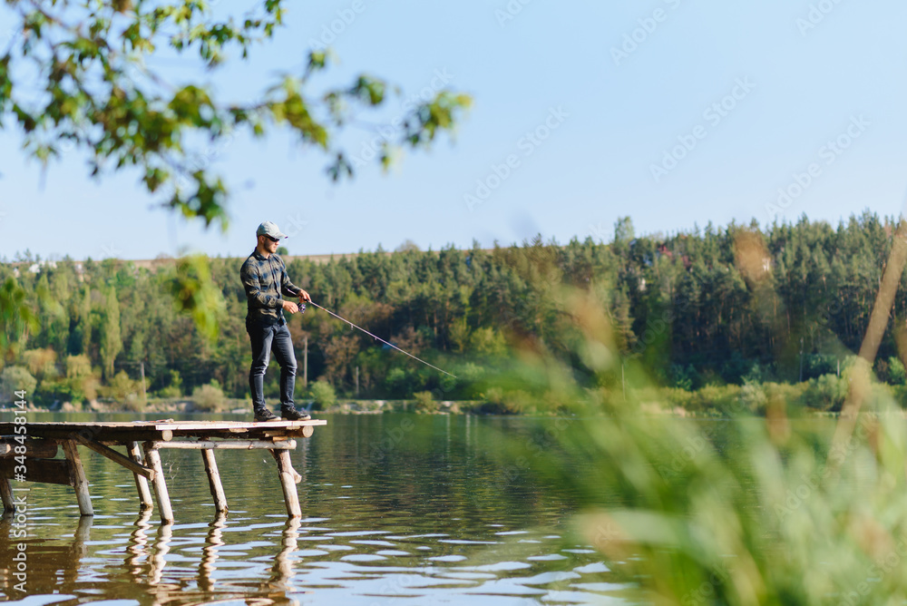 Fishing for pike, perch, carp. Fisherman with rod, spinning reel on river bank. Man catching fish, pulling rod while fishing on lake, pond with text space. Wild nature. The concept of rural getaway.
