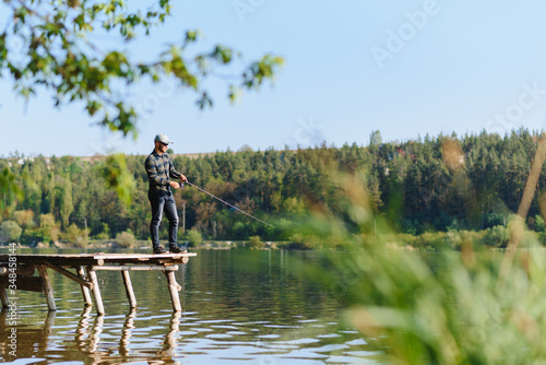 Fishing for pike, perch, carp. Fisherman with rod, spinning reel on river bank. Man catching fish, pulling rod while fishing on lake, pond with text space. Wild nature. The concept of rural getaway. © Serhii