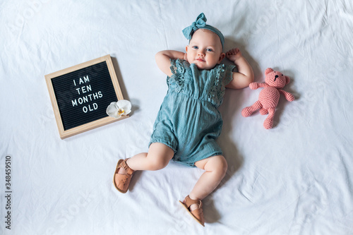 10 Ten months old baby girl laying down on white background with letter board and teddy bear. Flat lay composition.