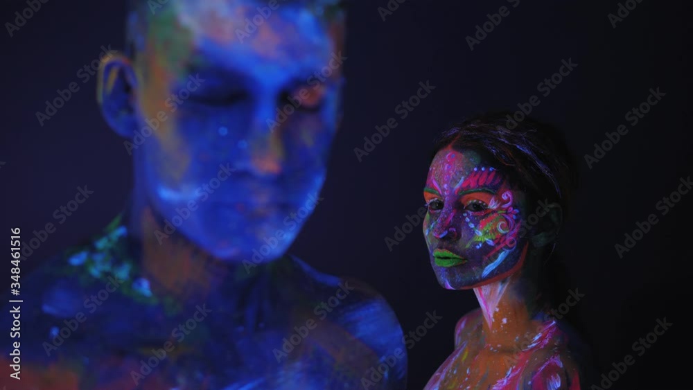 Portrait of a couple with UV drawings on the skin in the dark under the light of fluorescent lamps, the man in the foreground blurred. Body art on the couple's body glows in neon light. Portrait of a couple with UV drawings on the skin in the dark under the light of fluorescent lamps, the man in the foreground blurred. Body art on the couple's body glows in neon light.