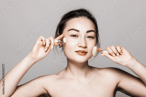 Portrait of happy young asian woman taking care of her skin and doing massage with cosmetology roller isolated on gray background