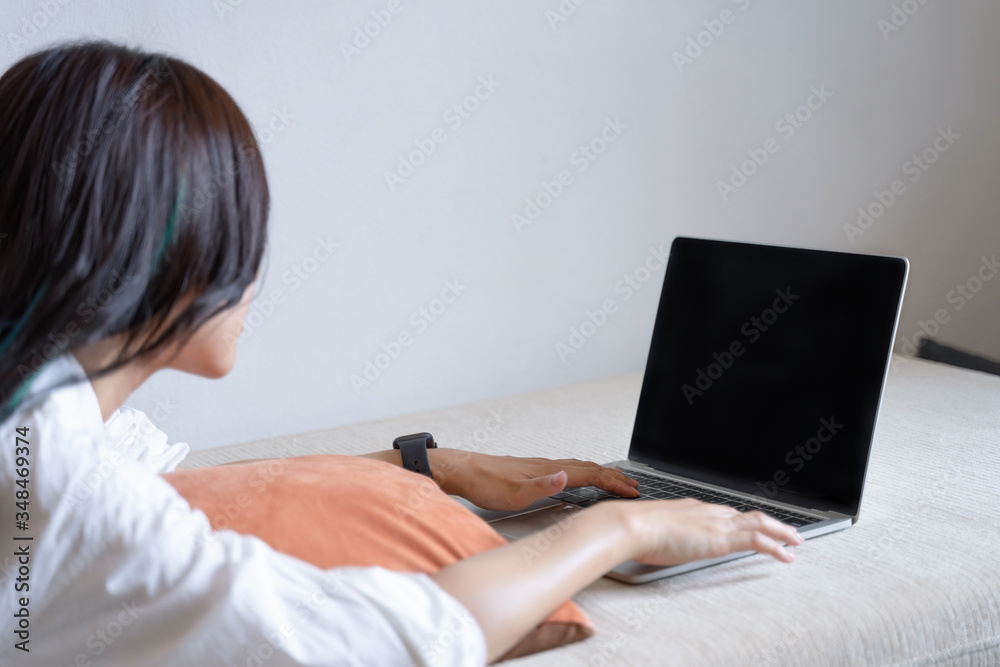 Fototapeta premium Back view of young female employee in casual wear typing on mockup black screen laptop in her home. Working from home concept